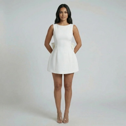 White fit-and-flare mini dress with pearl accents and cascading bow — front view styled with nude heels.