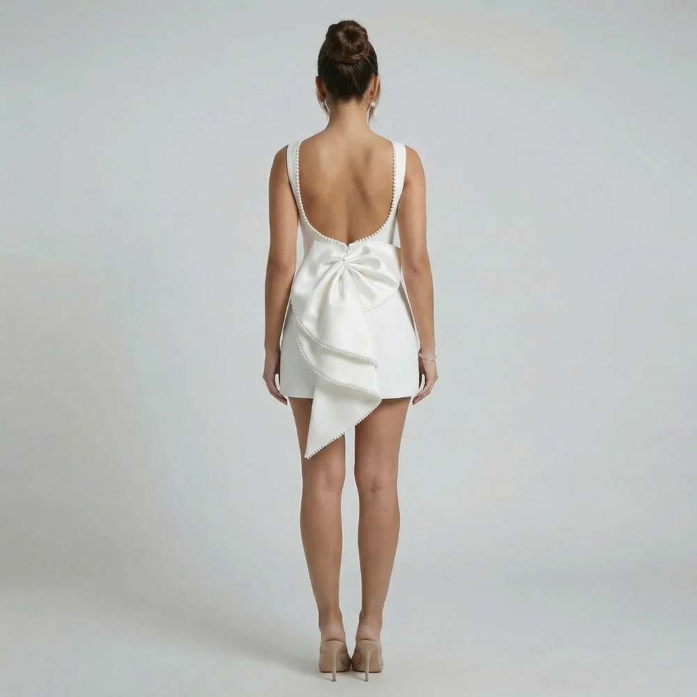 White fit-and-flare mini dress with low scoop back and elegant cascading bow — back view styled for an event.